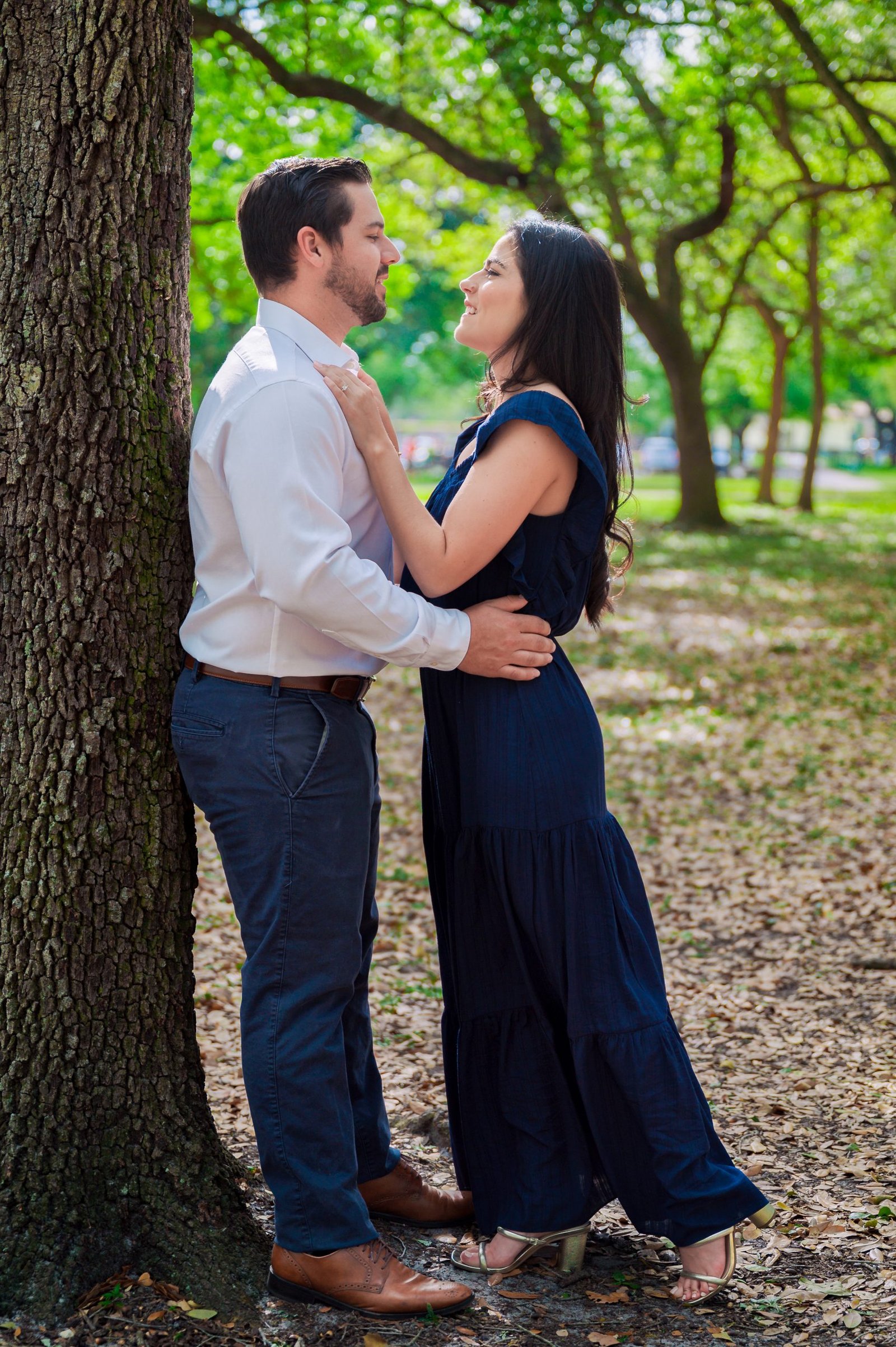 Engaged couple standing under a tree in a park, gazing into each other’s eyes