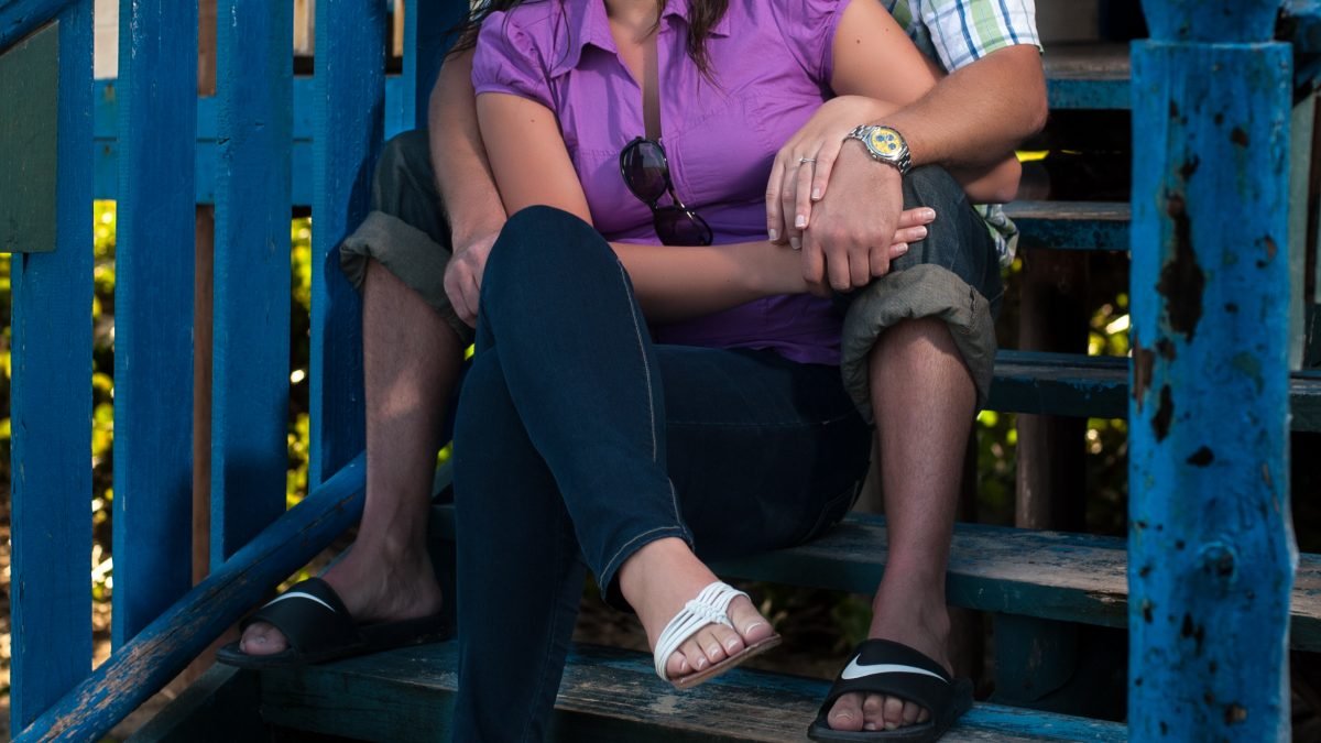 Engaged couple sitting on rustic blue wooden stairs of an old house, looking into the distance.