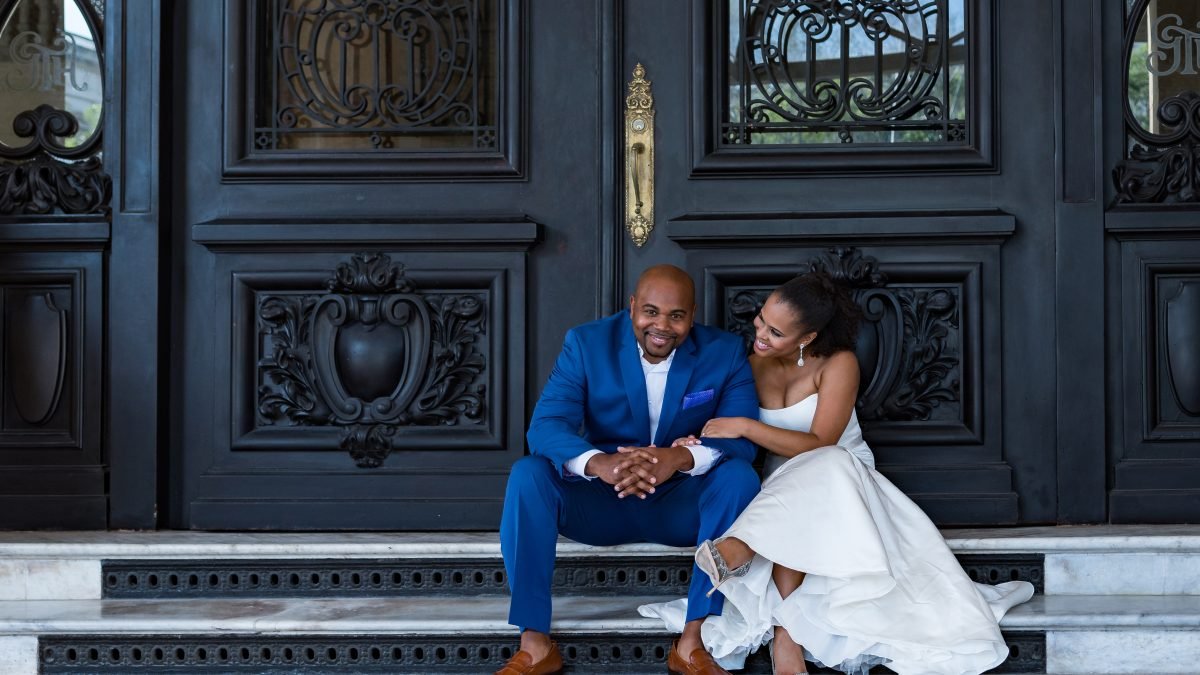 Bride lovingly gazing at groom while he looks at the camera, both seated on doorsteps in wedding attire