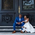Bride lovingly gazing at groom while he looks at the camera, both seated on doorsteps in wedding attire
