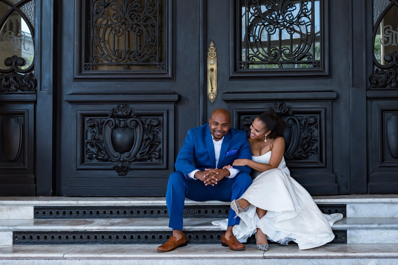 Bride lovingly gazing at groom while he looks at the camera, both seated on doorsteps in wedding attire