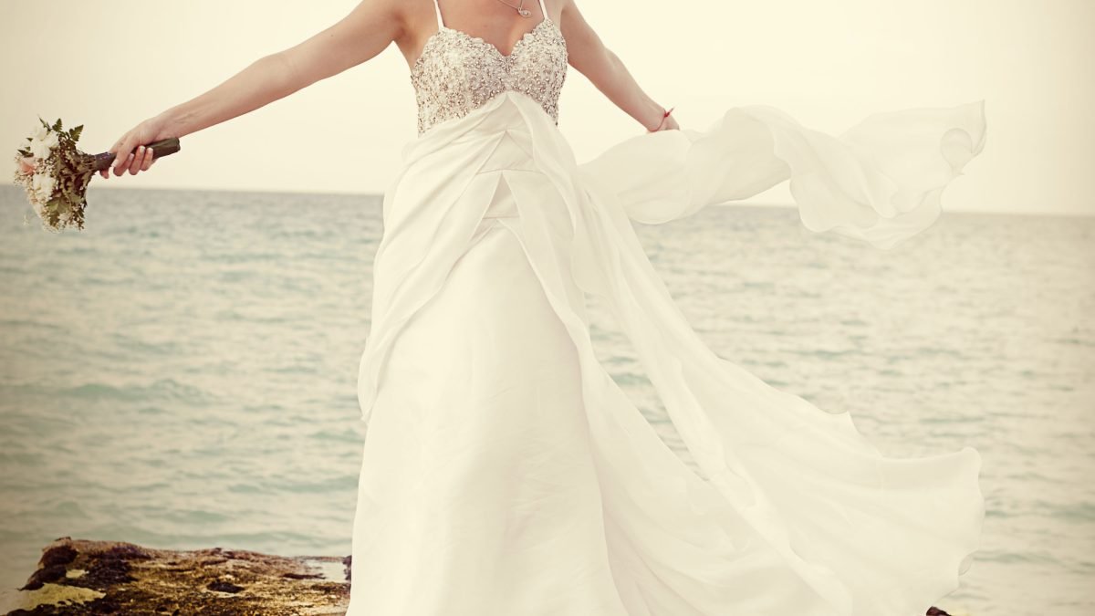 Bride spinning near the ocean, arms open wide, one holding a bouquet, the other stretching her dress to the side.