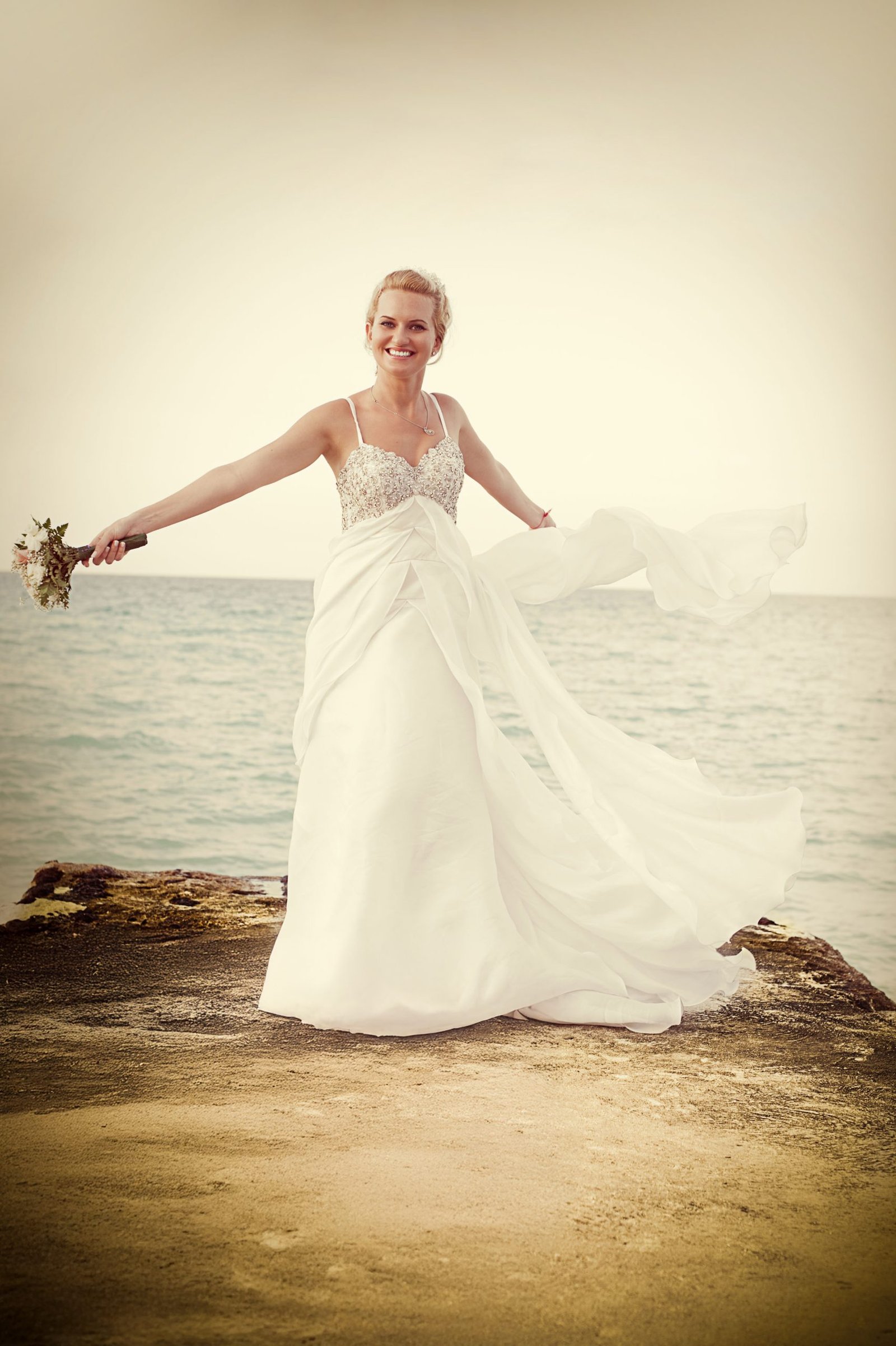 Bride spinning near the ocean, arms open wide, one holding a bouquet, the other stretching her dress to the side.
