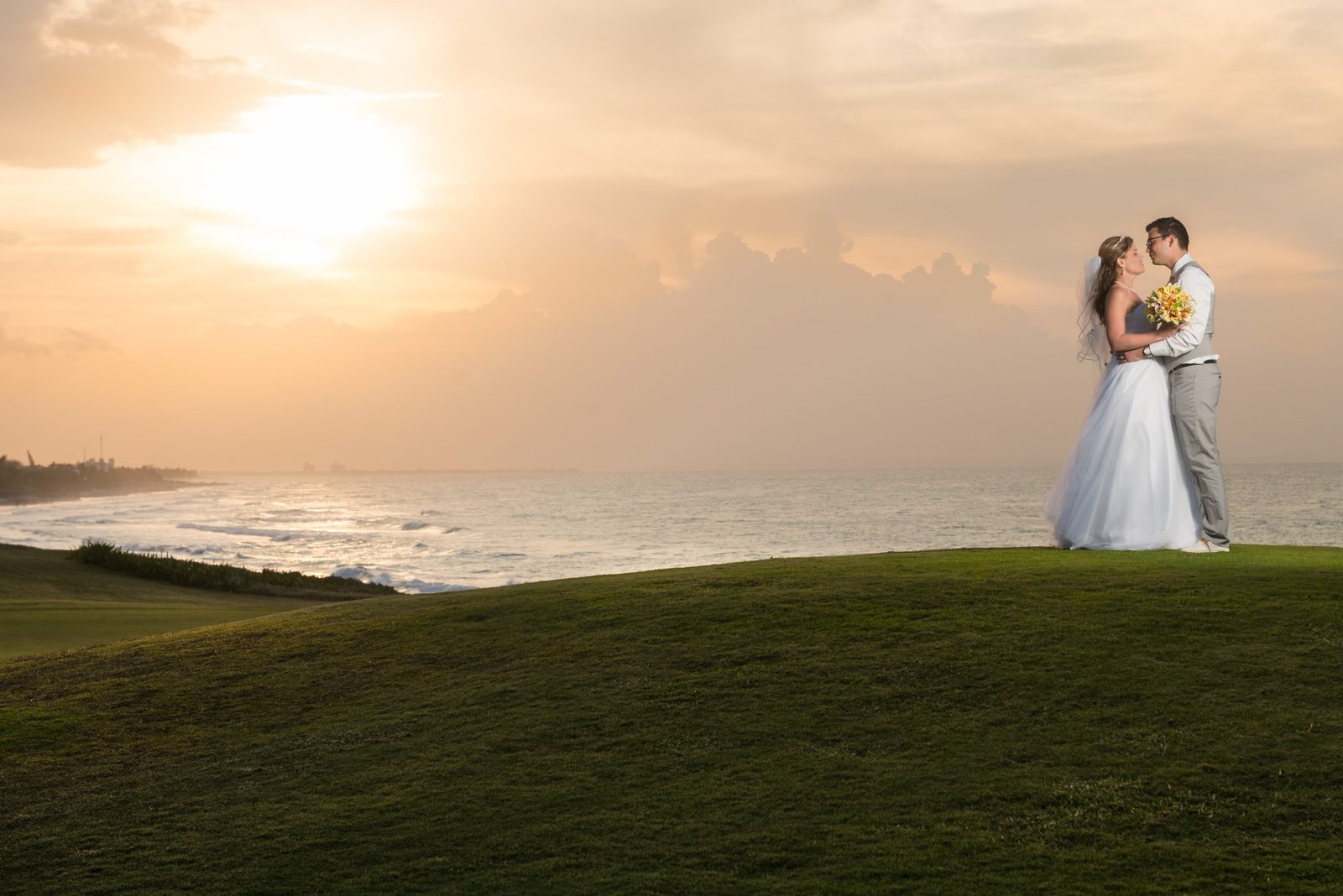 Wedding couple standing on top of a hill with ocean below and soft sun shining through a hazy sky.