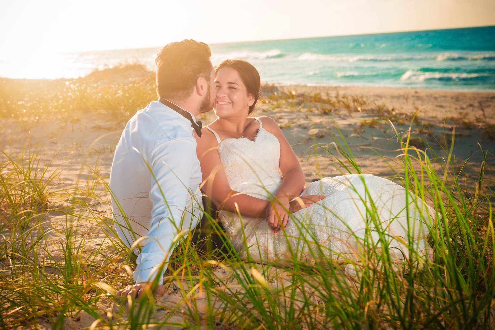 Wedding couple sitting on the sand with hazy sun and blue ocean in the background.