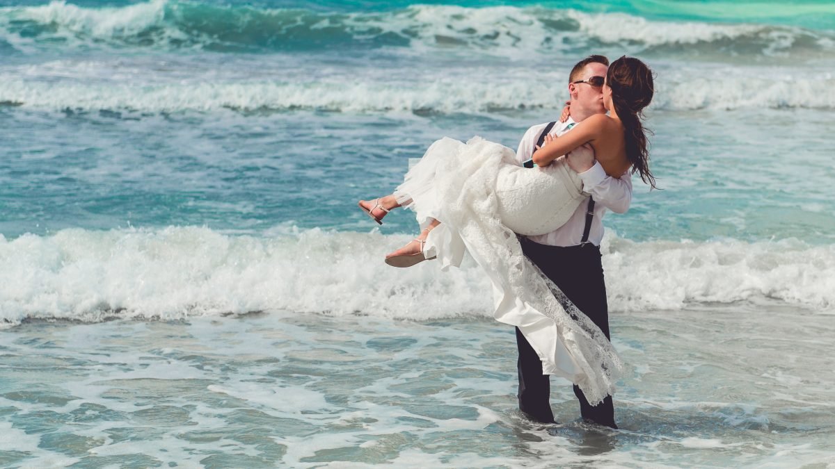 Groom carrying bride into the ocean during a beach wedding ceremony on a sunny day, captured by Alito Photography in South Florida.