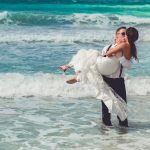 Groom carrying bride into the ocean during a beach wedding ceremony on a sunny day, captured by Alito Photography in South Florida.