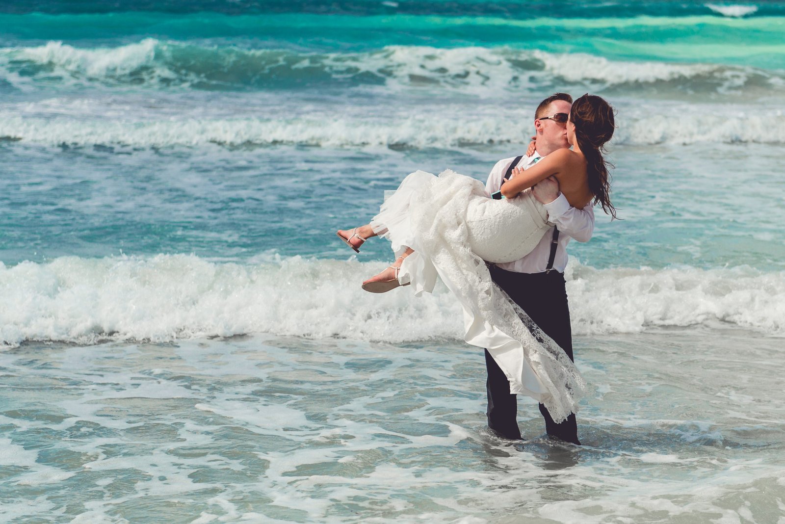 Groom carrying bride into the ocean during a beach wedding ceremony on a sunny day, captured by Alito Photography in South Florida.