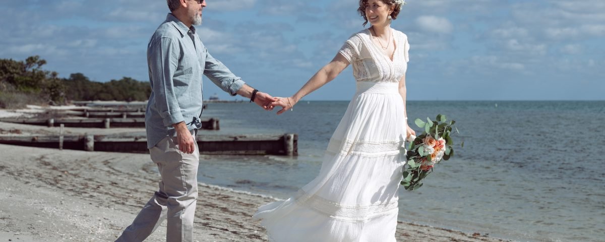 Newlywed couple walking by the ocean in Key Biscayne, Miami, with the bride leading her husband by the hand and smiling at him after their beach wedding ceremony.