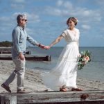 Newlywed couple walking by the ocean in Key Biscayne, Miami, with the bride leading her husband by the hand and smiling at him after their beach wedding ceremony.