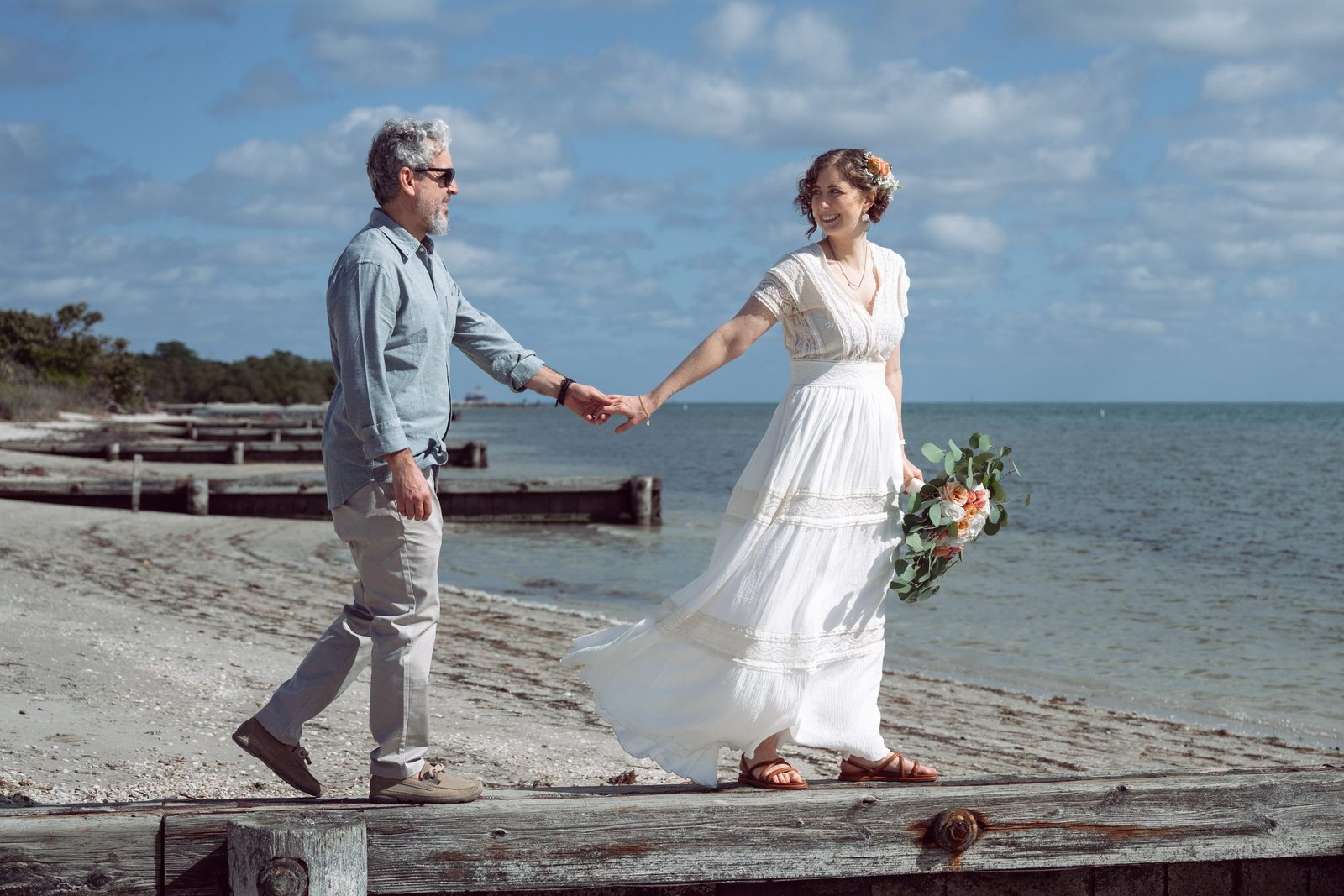Newlywed couple walking by the ocean in Key Biscayne, Miami, with the bride leading her husband by the hand and smiling at him after their beach wedding ceremony.